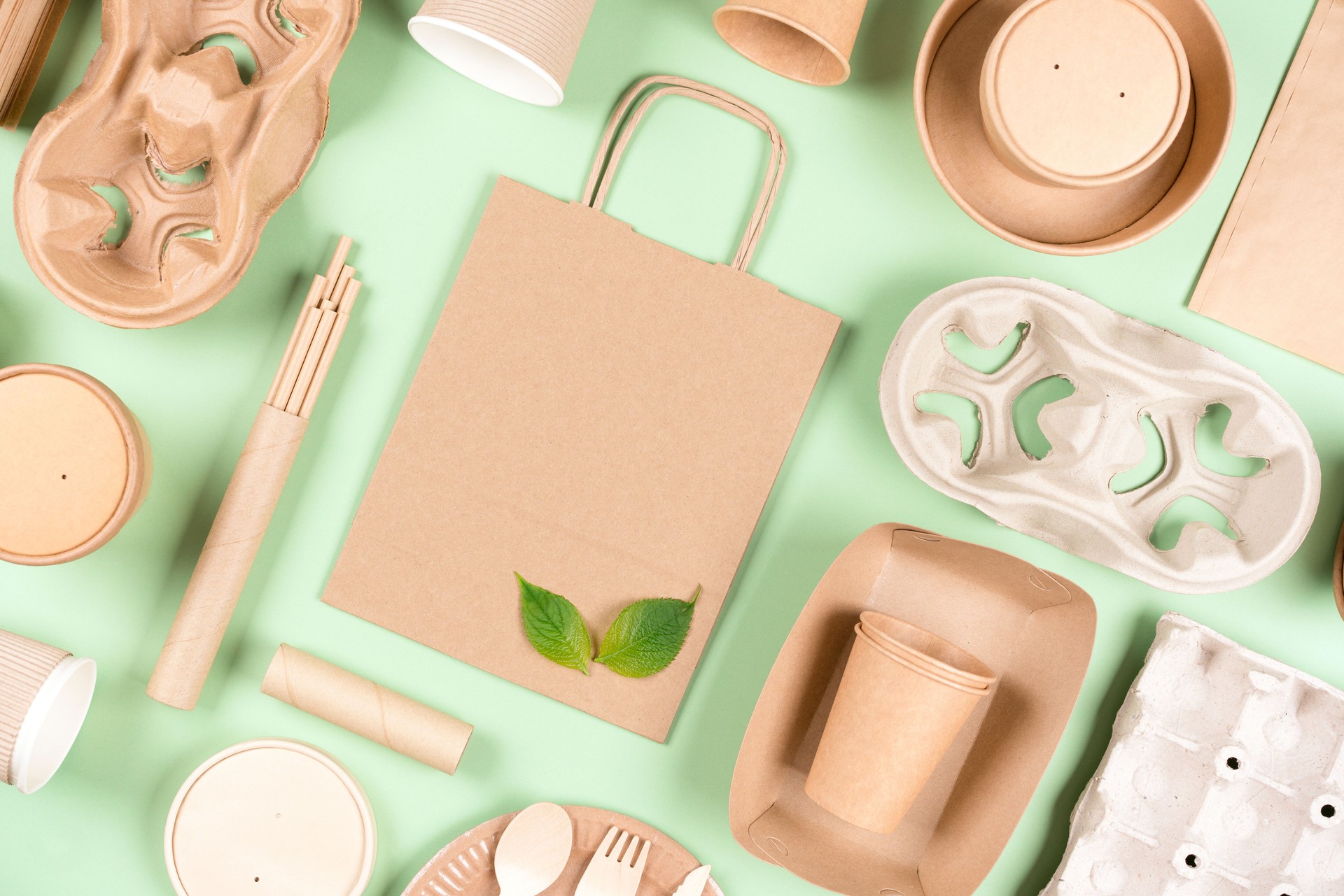 Paquete de utensilios de papel ecológico: recipientes de alimentos, bolsas de papel y portavasos, tazas de café sobre fondo verde claro. Concepto de envasado alimentario sostenible. Estilo laico de